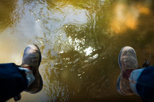 POV shot looking down at denim jeans and leather boots dangling over running water