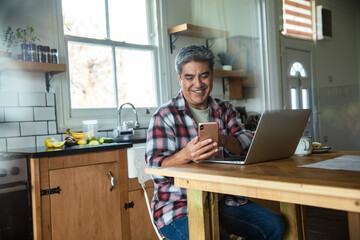 Smiling man using smartphone and laptop in home kitchen