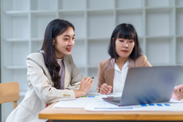 Obraz premium Asian businesswomen analyzing data and charts on a laptop, discussing work in a modern office environment