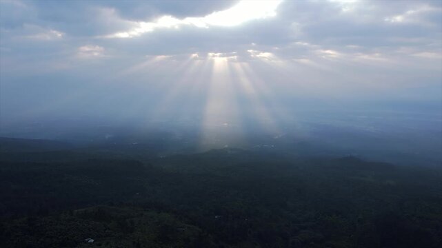 4K aerial view of beautiful dramatic sun rays breaking through clouds over a dark green forest hill.