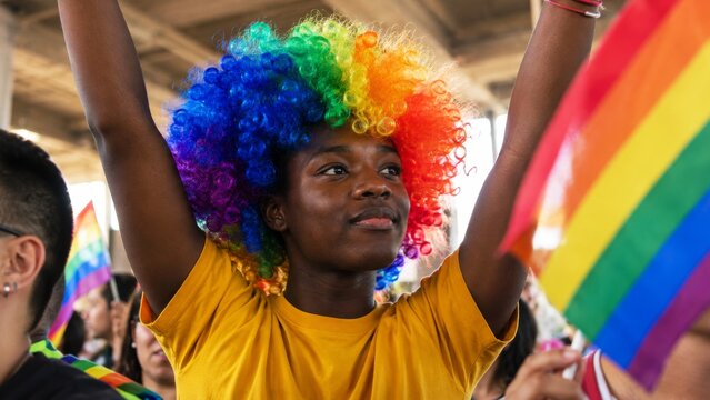 A joyful participant at a pride event, wearing a vibrant rainbow wig and raising their arms, celebrating diversity and inclusivity.