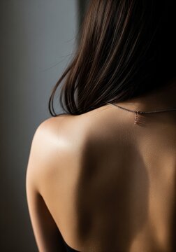 Close-up shot of a woman's bare back and shoulder from behind, showing smooth skin and soft lighting, with a delicate thin necklace on her neck