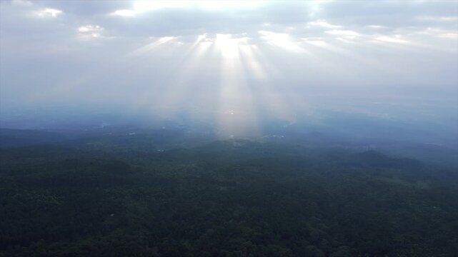 4K aerial view of beautiful dramatic sun rays breaking through clouds over a dark green forest hill.