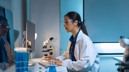 Female scientist wearing a lab coat and safety glasses, analyzing data on a computer in a modern research laboratory © Crystal