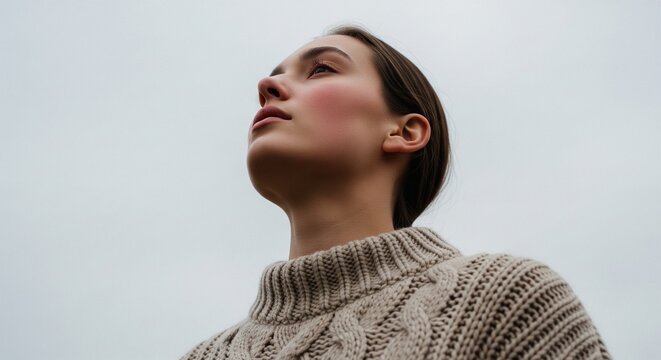 Low angle shot of a natural young woman looking up into a grey, overcast sky. She is wearing a heavy knitted beige sweater, creating a moody, peaceful autumn or winter atmosphere