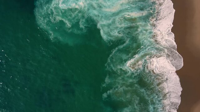 Aerial shot of the ocean with blue colours, waves doing  patters, rocks, sand and beach. Beautiful scenery