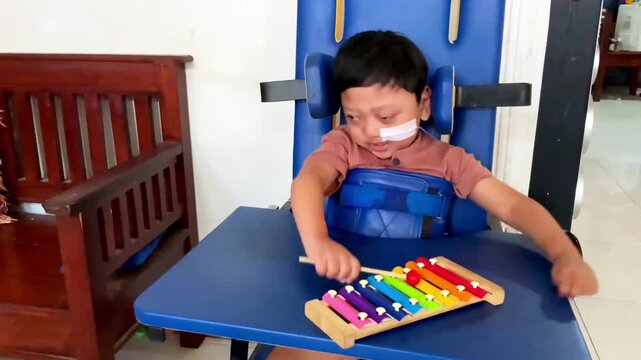 A young boy with a medical condition playing a colorful xylophone while sitting in a supportive chair