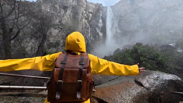 Happy female tourist wearing a bright yellow raincoat and backpack opens her arms wide, feeling the mist from the powerful bridalveil falls waterfall in yosemite national park, california