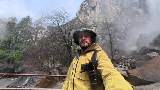 Happy male traveler with a camera and backpack filming himself on a trail at bridalveil fall in yosemite national park, gesturing towards the majestic waterfall during his adventure trip