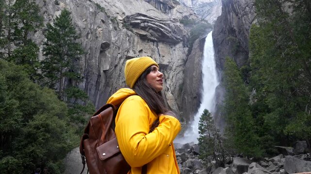 Young smiling woman with a yellow raincoat and backpack turning and looking at the camera with the amazing lower yosemite falls waterfall in the background, a popular landmark in california