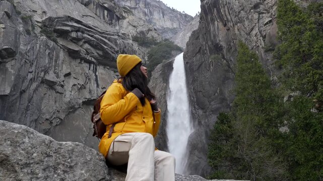 Happy female hiker in a bright yellow jacket sitting on a rock, admiring the stunning view of the powerful lower yosemite falls and looking at the camera, surrounded by granite cliffs