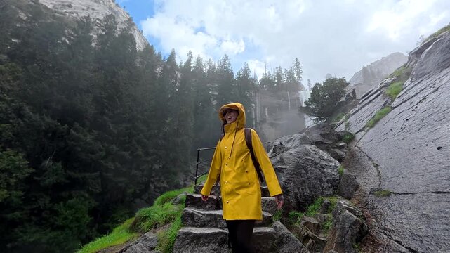 Young woman wearing a yellow raincoat and carrying a backpack walking up wet stone stairs on the mist trail, feeling the spray from vernal fall in yosemite national park, california