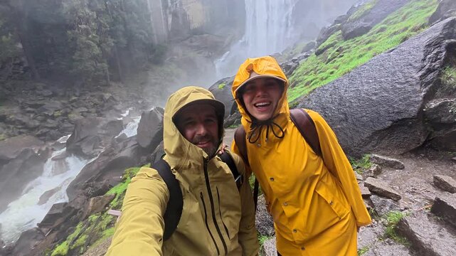 Cheerful young couple wearing raincoats and filming themselves on a smartphone while hiking the wet and misty trail next to a powerful waterfall in yosemite national park, california