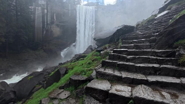 Powerful vernal fall creating a misty atmosphere along the wet stone staircase of the mist trail, a famous hiking path in california's yosemite national park surrounded by lush green nature