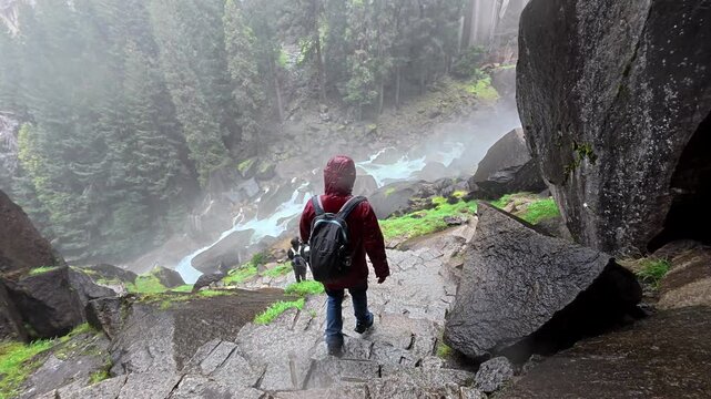 Young man wearing a red rain jacket and backpack carefully walking down the wet stone steps of the mist trail, surrounded by the powerful spray of a waterfall in yosemite national park