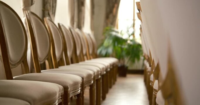 Cinematic historic mansion interior at sunset. Rows of antique chairs before concert. Large windows and a palm plant in the background. Warm evening light.