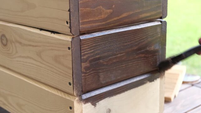 Brown oil for protection of wood by staining. Painting fence for protecting and pests. Woodworker applying finish to pine board box. Repairman varnishing with brush wooden fencing wall plank surface.