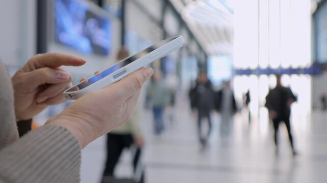 Hand holds tablet in busy transit hub. Person focused on screen, ignoring background movement. Blurred travelers walk past in hallway. Ideal for travel, technology, or modern commuting themes