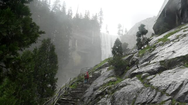 Lone hiker wearing a red raincoat walking up the wet stone steps of the famous mist trail, with the powerful vernal fall creating a dense spray in the majestic yosemite national park