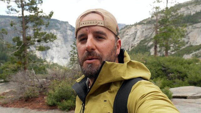 Young male influencer vlogging and taking a selfie video while hiking along the mist trail in yosemite national park, california, with nevada fall and vernal fall in the background