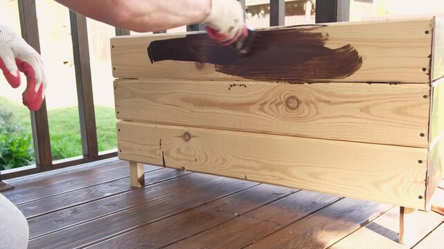 Brown oil for protection of wood by staining. Painting fence for protecting and pests. Woodworker applying finish to pine board box. Repairman varnishing with brush wooden fencing wall plank surface.