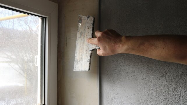 Interior finishing work, leveling a window slope. A man's hand holds a wide facade spatula and presses the blade of the spatula against the plane of the slope to the left of the wall corner to apply