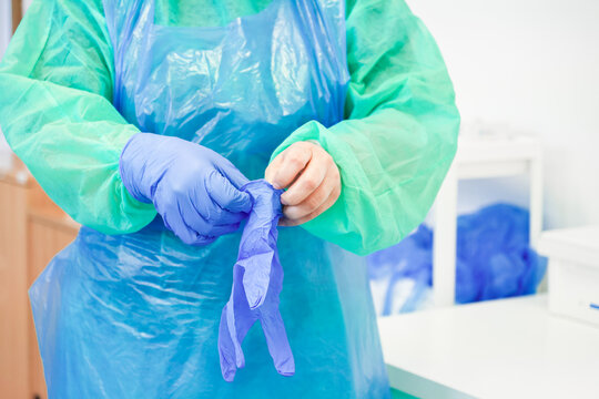 A medical professional puts on purple nitrile gloves while wearing a protective plastic apron and surgical gown to maintain strict sterility and hygiene in a clinical setting