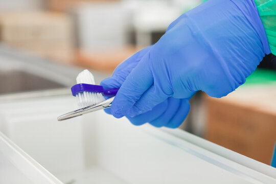 Gloved hand cleans a surgical instrument with a small scrubbing brush in a sterile hospital setting, representing the essential maintenance of medical equipment for patient safety
