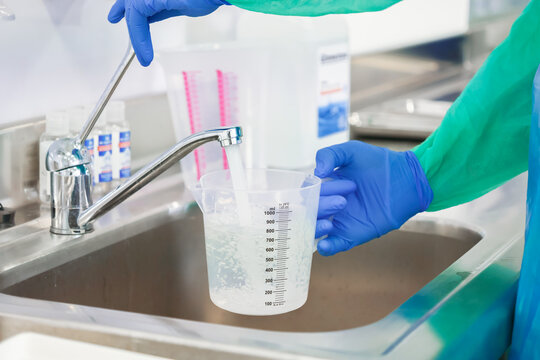 A laboratory worker in protective gear fills a plastic measuring cup with water at a stainless steel sink for precise liquid preparation tasks within a clean clinical facility