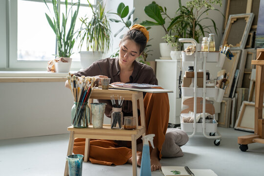 Pondering inspired woman painter enjoying creative process of creating new artworks at home gallery workshop, female artist sitting on floor drawing sketches, surrounded by art supplies and canvas