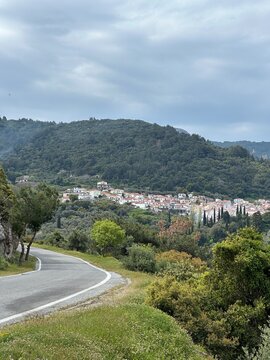 Road winds through green hills, leading to a small village in the distance under an overcast sky in the afternoon