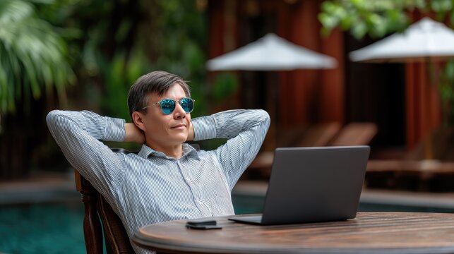 Man enjoys time by swimming pool with laptop and smartphone on table, smiling with relaxed pose in a sunny outdoor setting near a red building
