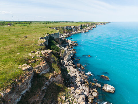 Aerial view of rugged rocky cliffs along the turquoise Black Sea coast with green meadows and wind turbines in the distance Kamen Bryag, Dobrich Province, Bulgaria.