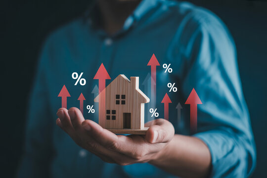 Person holding wooden house model with rising percentage symbols and arrows.