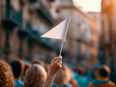 Hand holding white triangular flag raised above crowd at street demonstration during golden hour in urban setting