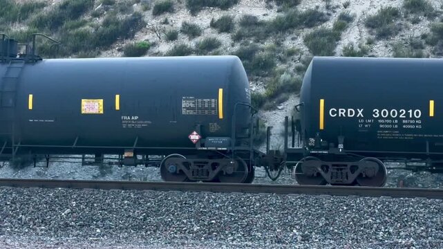 Two black tanker train cars move on railroad tracks with gravel below and sandy hills with green shrubs in the background on an overcast day in a remote outdoor setting