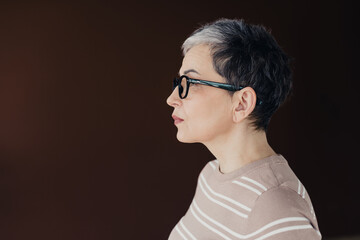 Profile portrait of a thoughtful middle-aged woman with short gray hair and glasses against a dark background © deagreez