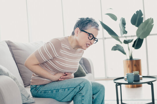 Senior woman clutching stomach in pain while sitting on sofa at home, experiencing abdominal discomfort and distress during digestive or menstrual cramp episode