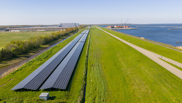 Aerial view of the ENGIE Maxima plant solar farm with long rows of panels next to a green dike and industrial port in Lelystad, Flevoland, Netherlands.