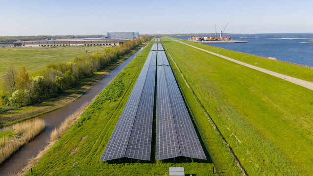 Aerial view of the Maxima plant solar farm featuring long rows of photovoltaic panels on a green dike beside a canal in Lelystad, Flevoland, Netherlands.