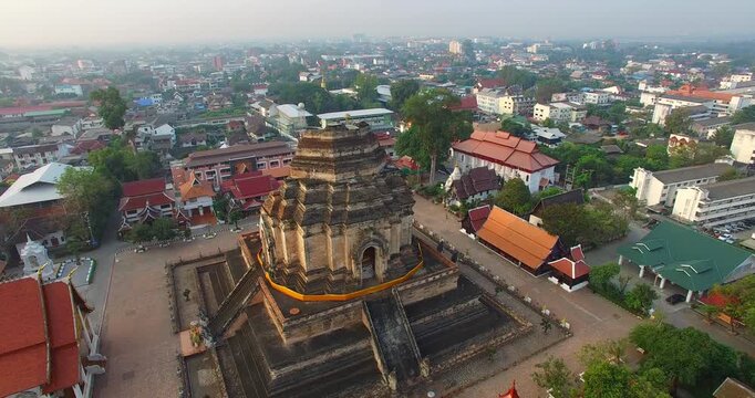 High angle drone shot capturing the grandeur of Wat Chedi Luang temple with its towering ancient stupa and beautifully crafted serpent statues along the stairway. Surrounded by old city walls
