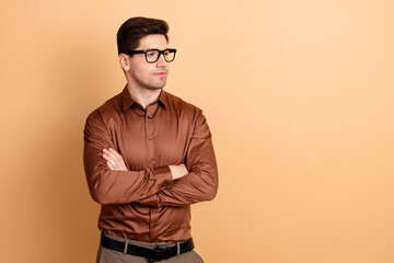 Obraz premium Confident young man in glasses and brown shirt posing with arms crossed looking thoughtful and professional against a warm neutral background