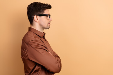 Young man in brown shirt and glasses looking sideways in profile on beige background, thoughtful studio portrait with arms crossed and neutral expression © deagreez