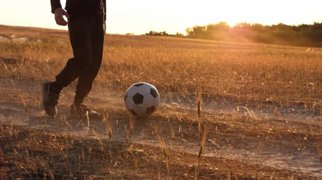Close-up of boys legs kicking soccer ball on rural field at sunset, dynamic movement in warm golden light. Concept of football training, skill development and active lifestyle grassroots sport. Child