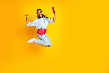Obraz premium Joyful young woman jumps in the air with yellow background while taking a selfie to promote fashion lifestyle and fun