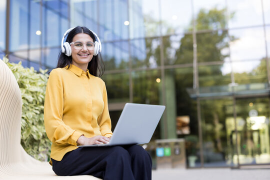 Young woman smiling, wearing headphones and clear glasses, working or studying on a laptop while sitting on a modern bench outdoors in front of a glass building