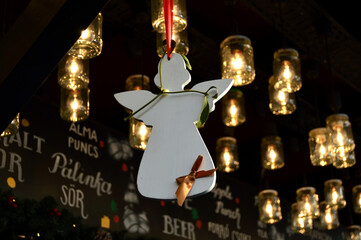 Obraz premium Christmas market scene, angel ornament hanging, warm festive lights, cozy winter atmosphere, European holiday fair, seasonal street decoration, gift shopping mood, evening bokeh glow. 