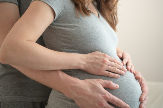 Pregnant woman with partner placing hands over her belly in shared intimate moment. Expecting mother and husband embracing from side in studio with neutral background. Mom-to-be celebrating shared