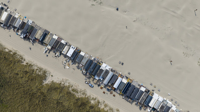 Aerial view of a long row of small beach houses lined up on a sandy shore next to grassy dunes in Velsen-Noord, North Holland, Netherlands.