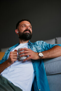 Smiling adult relaxing by the sofa with a coffee mug and wireless earbuds while listening to music or a podcast